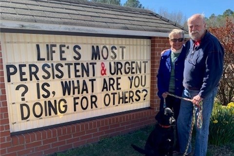 Man and woman with dog stand beside a sign about volunteering